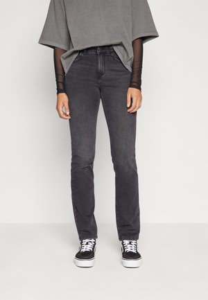 Personne portant un jean skinny noir, des manches longues en maille noire sous un t-shirt gris oversize, et des baskets noires avec des lacets et semelles blanches.