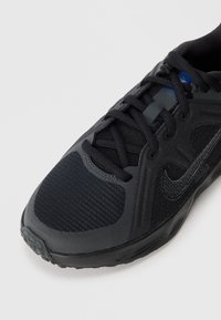 Ténis desportivo preto com parte superior texturizada, painéis em malha e um logótipo preto brilhante. Apresenta atacadores e uma sola de borracha com padrão antiderrapante.