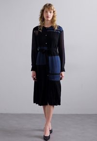 Črna obleka z dolgimi rokavi, gumbnim zapenjanjem spredaj, nabrano krilo in mešanico modrih vzorčenih panelov. Styled with a tie belt and black shoes.