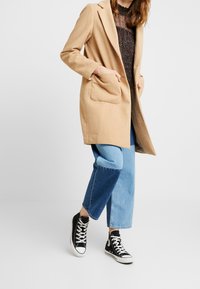 Cappotto in misto lana beige con design sartoriale, due tasche frontali, indossato sopra un top nero con fantasia e abbinato a jeans blu cropped e sneakers nere.