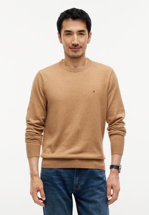 Homme portant un pull beige à col rond et un jean bleu, debout devant un fond blanc uni, avec une montre noire au poignet gauche.