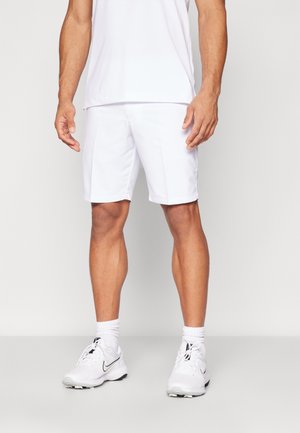 Mężczyzna ubrany w białe sportowe szorty, białą koszulkę z krótkim rękawem, białe skarpety i białe buty sportowe marki Nike, stojący na jednolitym tle.