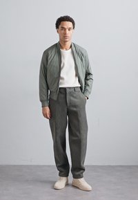 Ljusgrön bomberjacka över en vit strukturerad skjorta, tillsammans med lösa mörkgröna byxor och beige sneakers. Minimalistisk design.