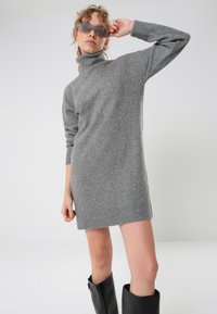 Gray knit sweater dress with a high turtleneck and ribbed hem. Paired with dark sunglasses and black knee-high boots.