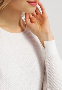 Bílý pletený svetr s texturovaným vzorem včelí plástve, žebrovaným límcem a manžetami. Materiál vypadá měkce a lehce.