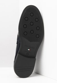 Suela de zapato de goma negra con patrones de agarre circulares, nombre de marca embosado "Nero Giardini" y detalle de la bandera italiana. Forma suave y curvada.