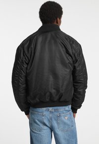 Jachetă bomber neagră cu un finisaj lucios, guler și manșete ribbed, având un tiv elastic. Pur tată în fața unei perechi de blugi din denim de culoare albastru deschis.