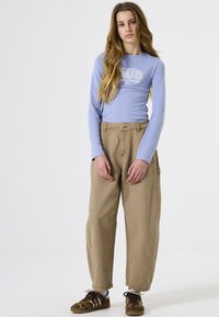 Lichtblauwe overhemd met lange mouwen en witte tekst, ruime beige broek, bruine schoenen met patronen en veters, casual outfit in een neutrale setting.