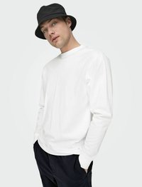 Wit longsleeve shirt met een ronde hals, gecombineerd met een zwarte bucket hat. Het model staat met de handen in de zakken, gekleed in donkere broek.