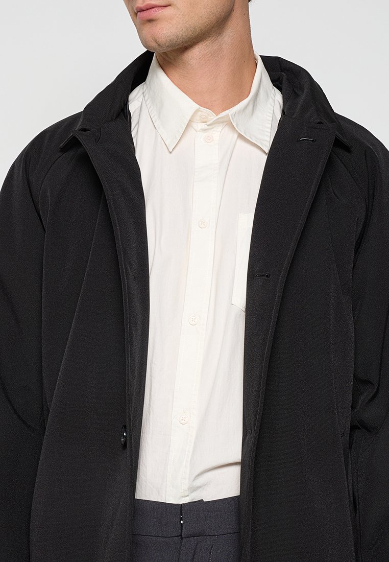 Zwarte buitenkleding met een gladde textuur, met een ontspannen pasvorm en open kraag, gedragen over een wit overhemd met knopen en een borstzak.