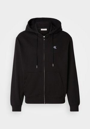 Zwarte zip-up hoodie gemaakt van zachte stof. Heeft een verstelbare capuchon, voorzakken en een klein geborduurd logo op de borst.