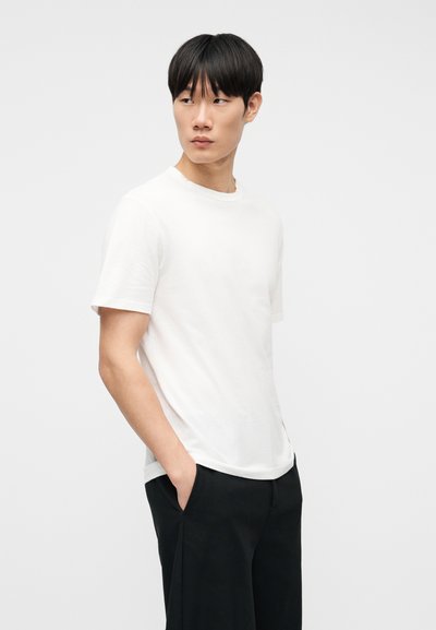 Άνδρας που φοράει λευκό απλό κοντομάνικο μπλουζάκι και μαύρο παντελόνι, με τα χέρια στις τσέπες, κοιτάζοντας προς τα πλάγια, μπροστά σε απλό φωτεινό φόντο.