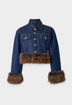 Femme portant une veste et un pantalon en denim bleu foncé avec une bordure en fourrure marron aux poignets, à la taille et sur les jambes, assortis à des talons ouverts noirs.