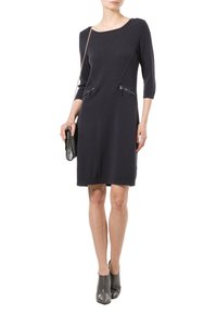 Abito a maglia colore navy con maniche a tre quarti, dotato di due tasche con zip. La modella tiene in mano una borsa metallica piccola e indossa stivaletti grigi alla caviglia.
