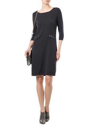 Robe tricotée navy avec des manches trois-quarts, dotée de deux poches zippées. Le mannequin tient un petit sac métallique et porte des bottines grises.