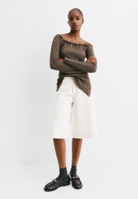 Top marrone off-shoulder in tessuto testurizzato, pantaloni corti bianchi a gamba larga e mocassini scuri con fibbia. La superficie liscia del tessuto e i colori a contrasto creano un interesse visivo.