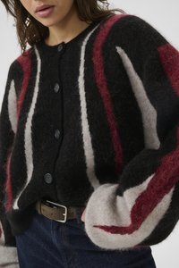 Frau in einem flauschigen schwarzen Cardigan mit weißen und roten vertikalen Streifen, dunklen Jeans und einem braunen Gürtel, mit einer Hand in der Tasche.