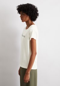 T-shirt crème clair avec un col rond et des manches courtes ; arbore du texte noir sur la poitrine. Associé à un bas vert olive.