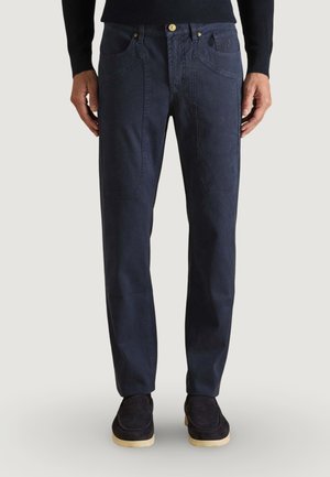 Mand iført marineblå bukser med lige ben og store frontlommer, sort sweater og sorte slip-on sko med beige såler.