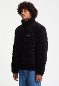 Homme aux cheveux bouclés portant une veste matelassée noire et un jean bleu, regardant vers la gauche sur un fond blanc uni.