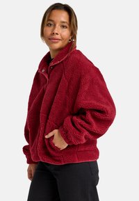 Pullover in pile rosso con colletto alto, dotato di chiusura a scatto, vestibilità oversize e tasche frontali. Tessuto morbido con un motivo trapuntato.