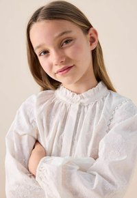 White embroidered blouse with ruffled collar and long puff sleeves, featuring delicate floral patterns throughout the fabric.