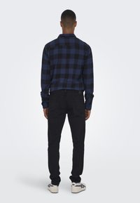 Zwart en blauw geruite flanellen overhemd met lange mouwen, gecombineerd met zwarte jeans en witte sneakers. Uitzicht van de achterkant, waarbij de pasvorm wordt getoond.