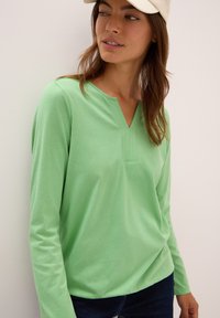 Frau in einem hellgrünen Langarmshirt mit einem V-ausschnitt und einer weißen Kappe, stehend vor einem schlichten weißen Hintergrund.