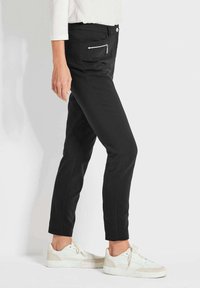 Schwarze Stretchhosen mit schlanker Passform, ausgestattet mit einer seitlichen Reißverschlusstasche und minimalen Nähdetails, kombiniert mit weißen Sneakers.