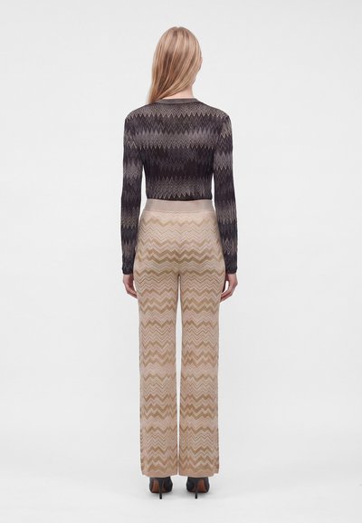 Femme debout de dos portant un pull en zigzag noir et gris et un pantalon large beige à motif zigzag avec des talons noirs.