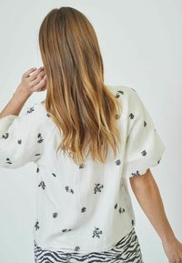 Blusa bianca con ricami floreali neri, vestibilità ampia, tessuto texturizzato e maniche corte; abbinata a una gonna a righe bianco e nera.