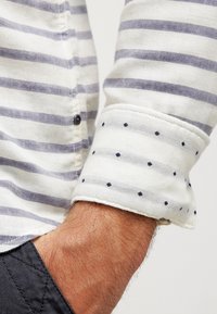 Camicia con colletto caratterizzata da righe orizzontali blu e bianche, con un polsino a pois in tessuto leggero a contrasto. Dettagli con bottoni visibili.