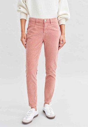Femme portant un pantalon en velours côtelé rose taille haute, coupe slim, avec poches avant et baskets blanches, debout devant un fond uni.