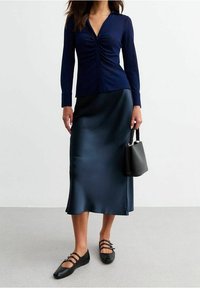 Marineblaues, geripptes Oberteil mit Knopfdetails und geraffter Vorderseite, kombiniert mit einem glatten, satinartigen dunkelblauen Midirock und schwarzen flachen Schuhen. Schwarze Handtasche inklusive.