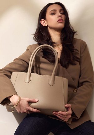 Beige Leder-Shopper mit strukturierter Oberfläche, zwei schlanken Griffen und goldfarbener Markenlogo. Getragen mit einem braunen Blazer und dunklen Jeans.