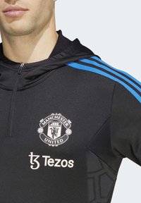 Felpa nera del Manchester United con chiusura a zip, strisce di accento blu e un logo del club in evidenza con il marchio "Tezos" sotto.