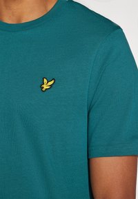 T-shirt turchese a maniche corte realizzato in morbido tessuto con scollatura rotonda, caratterizzato da un piccolo logo di un uccellino giallo ricamato sul petto.