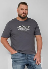 Grijze crewneck t-shirt van katoen, met op de voorkant in witte letters de tekst "Avontuurlijke Gevoelens Vooruit! Abonneer je op de Natuur." Zichtbare spijkerbroek.