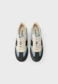 Zilā, melnā un bēšā krāsā krāsotu zābaku pāris ar zamša tekstūru, skatīts no augšas uz vienkārša balta fona.