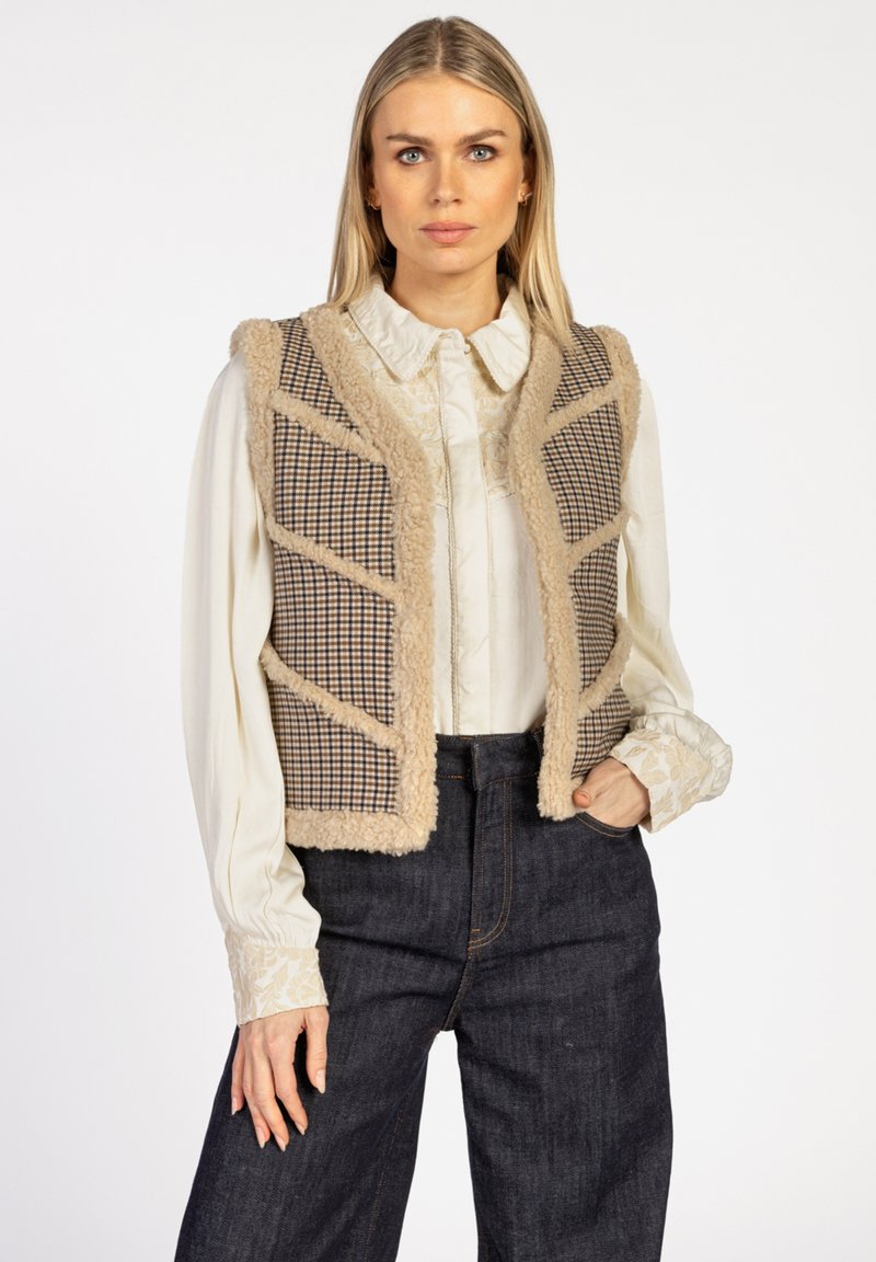 Beige geruiten vest met een pluizige rand, met een geometrisch patroon, gedragen over een lichtblouse met ingewikkelde mouwen en donkerdenim broek.