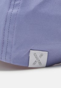 Lila Kappe aus Stoff mit einer strukturierten Oberfläche. Hat ein weißes Etikett mit einem metallischen silbernen "X"-Logo. Sichtbare Nähdetails.