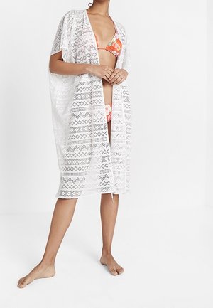 Vrouw draagt een oranje bikiniset met bloemenprint en een knielange, witte gehaakte open voorkant overgooi, staat blootsvoets op een witte achtergrond.