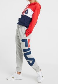 Röd och marinblå färgblockerad huvtröja med vita accenter, parat med grå sweatpants med en stor blå Fila-logotyp. Vita sportiga sneakers.