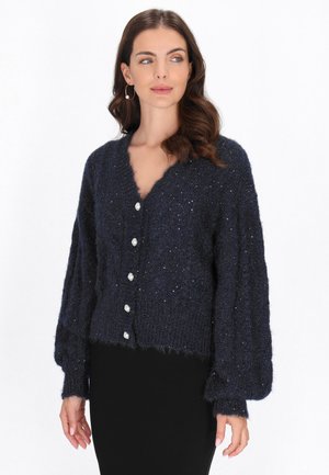 Jonge vrouw met golvend bruin haar, die een navyblauw vest met parelknopen en opvallende oorringen draagt.