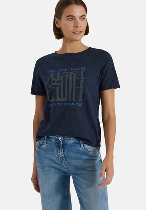 Femme portant un t-shirt bleu marine avec un texte en pointillés "TAKE MORE CHANCES DANCE MORE DANCES" et un jean bleu clair, regardant légèrement à droite.