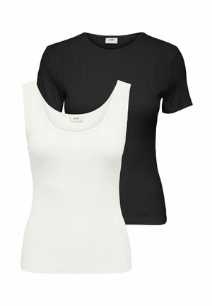 Femme debout portant un haut noir près du corps à manches courtes, un pantalon noir à jambes larges et des chaussures blanches pointues sur un fond uni.