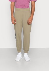 Beige sweatpants med elastisk midjeband och benslut, med en tryckt logodetalj på vänster ben. Bärs med en rosa t-shirt och vita sneakers.