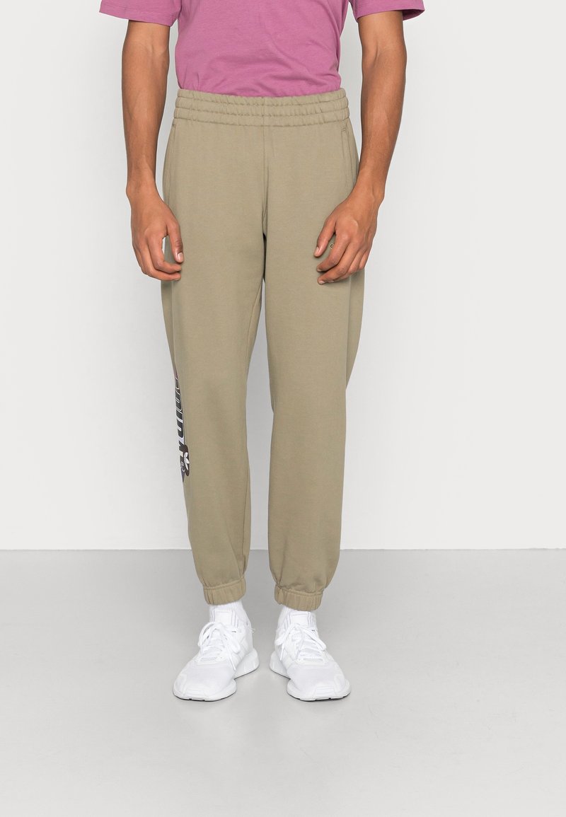 Beige sweatpants med elastisk midjeband och benslut, med en tryckt logodetalj på vänster ben. Bärs med en rosa t-shirt och vita sneakers.
