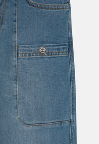 Doppio petto di jeans in azzurro chiaro con una tasca frontale e cuciture arancioni; presenta un bottone metallico rotondo e con marchio sulla tasca superiore.