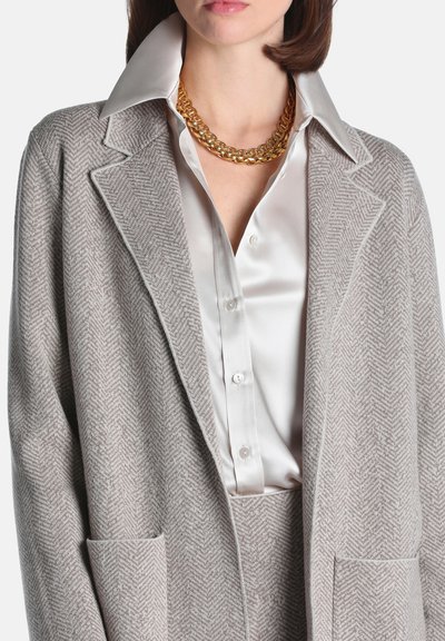 Femme portant un blazer gris clair à motif chevrons, pantalon assorti, chemise blanche en satin boutonnée et un collier chaîne épais en or.